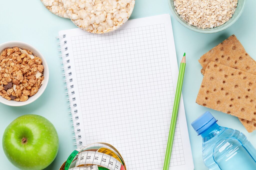 Blocco appunti a quadretti con matita verde, mela, metro da sarta, bottiglia d'acqua e cereali per piano nutrizionale e dieta personalizzata.